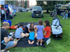 EMS officer showing children and adults how to do CPR on dummies on blankets on the ground, in grassy park area