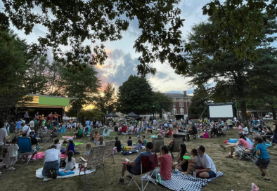 image of many people sitting on blankets on the village green looking at a stage and movie screen