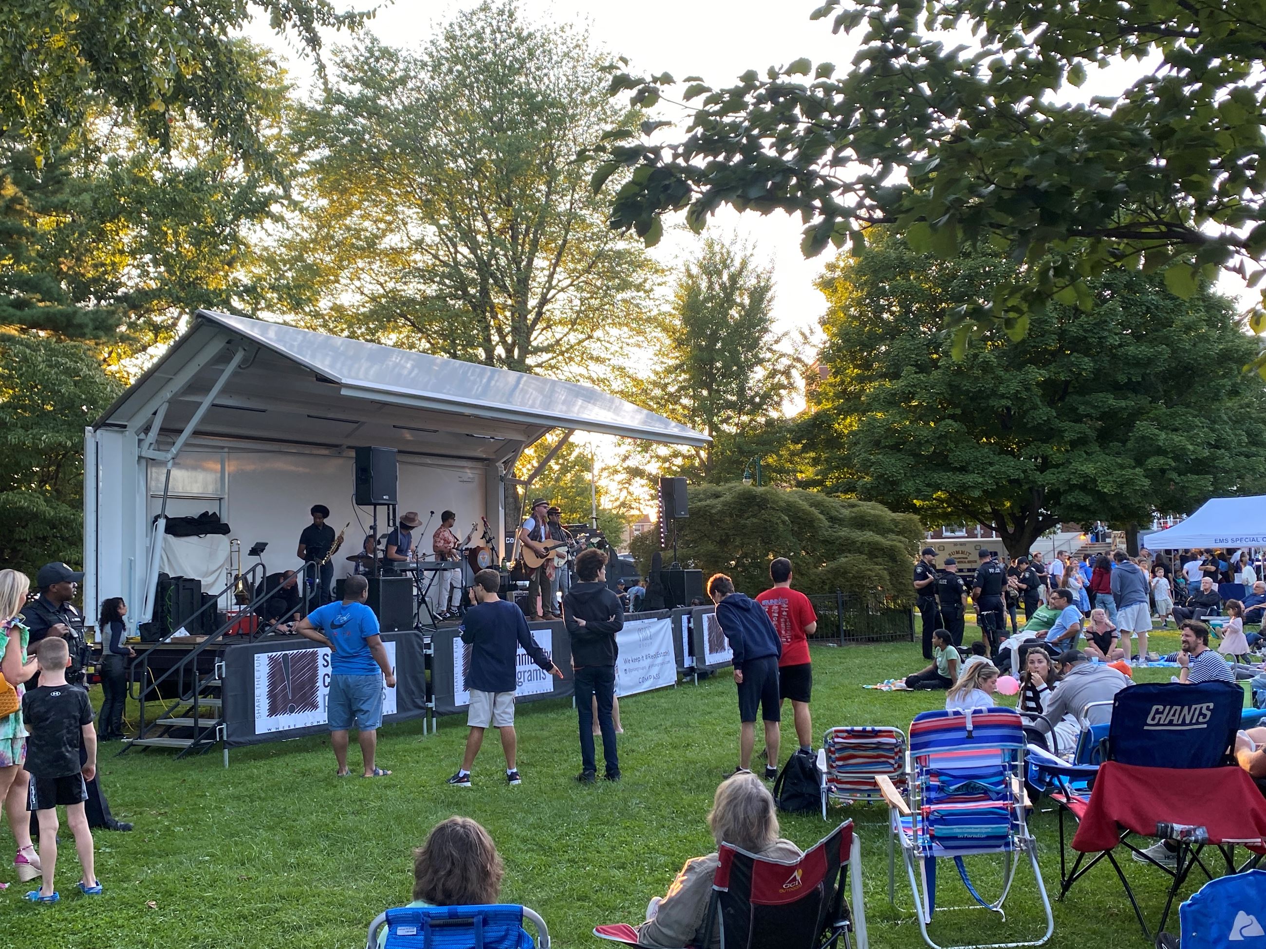 Large crowd of people seated in lawn chairs and blankets in grassy park area watching concert on stage