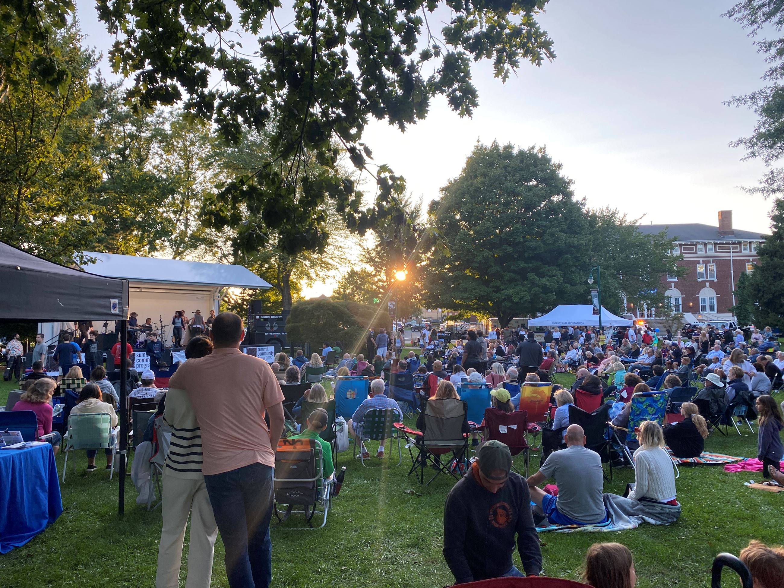 Large crowd of people seated in lawn chairs and blankets in grassy park area watching concert on stage