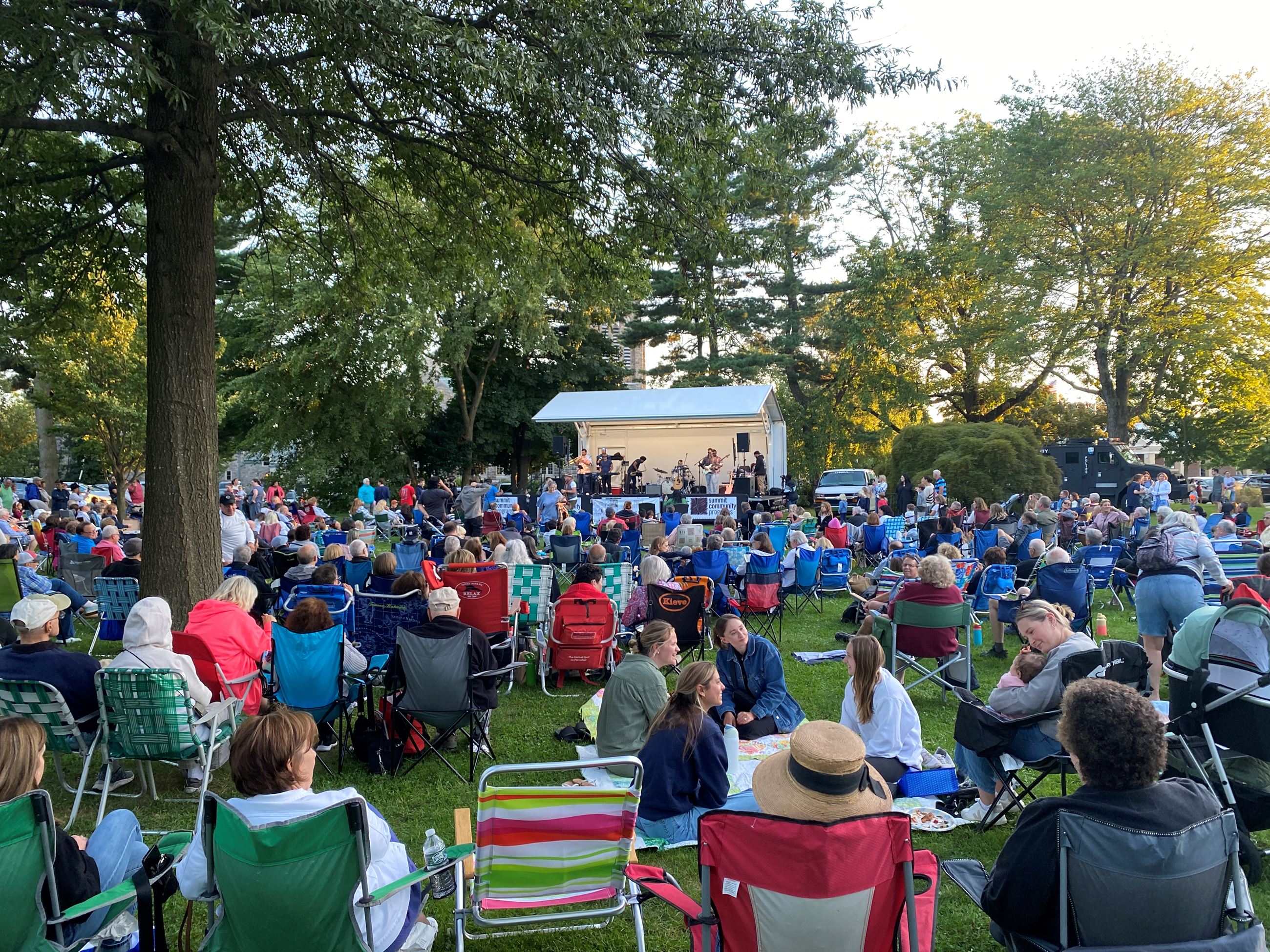 Large crowd of people seated in lawn chairs and blankets in grassy park area watching concert on stage