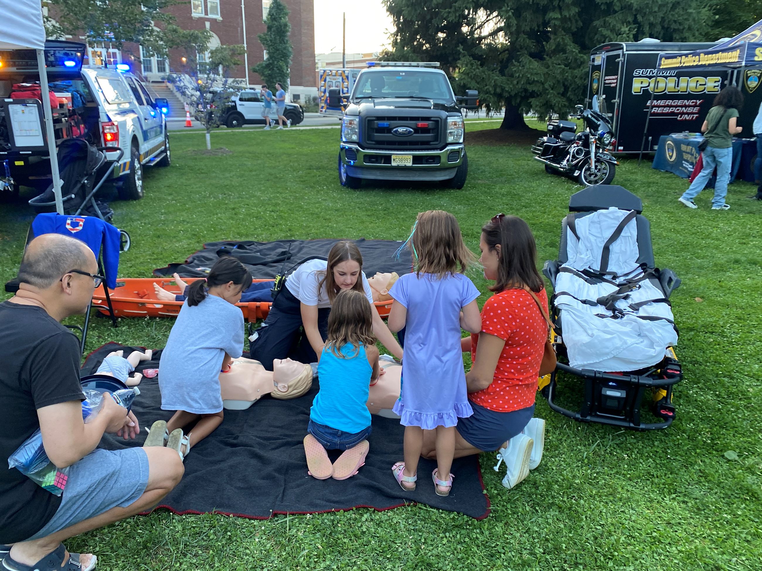 EMS officer showing children and adults how to do CPR on dummies on blankets on the ground, in grassy park area