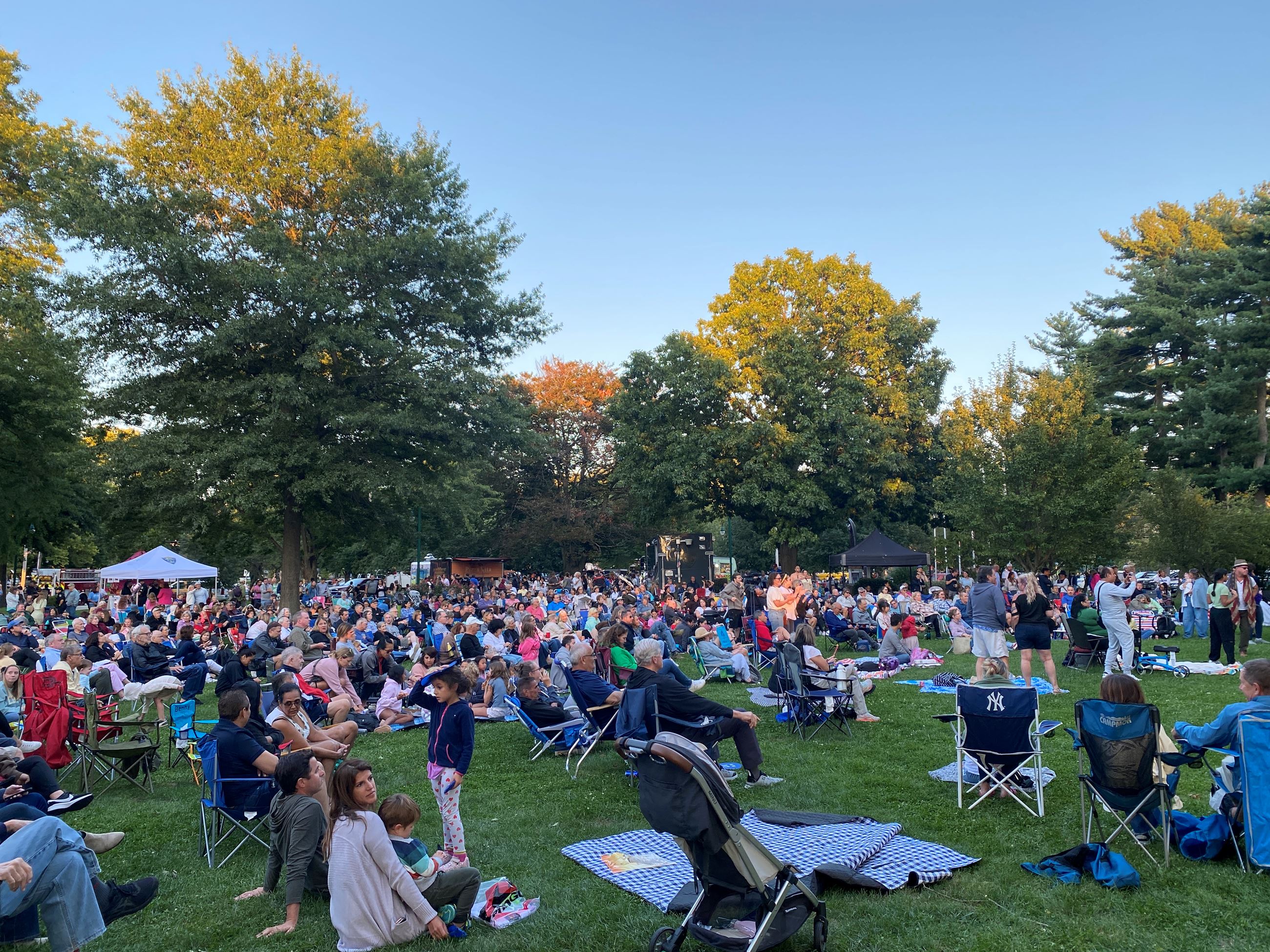 Large crowd of people seated in lawn chairs and blankets in grassy park area watching concert on stage